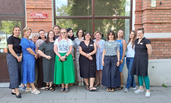 Grupa kilkunastu kobiet stojących przed wejściem do ceglanego budynku, na którym przyczepiona jest czerwona tabliczka z białym napisem "Wolskie Centrum Kultury"