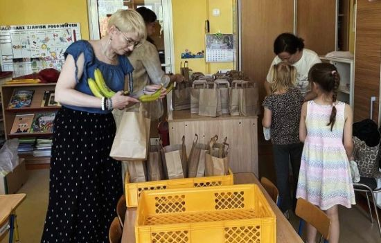 Dzieci i dorośli pakują kanapki do papierowych toreb, na pierwszym planie stoją żółte plastikowe skrzynki, w tle sala przedszkolna