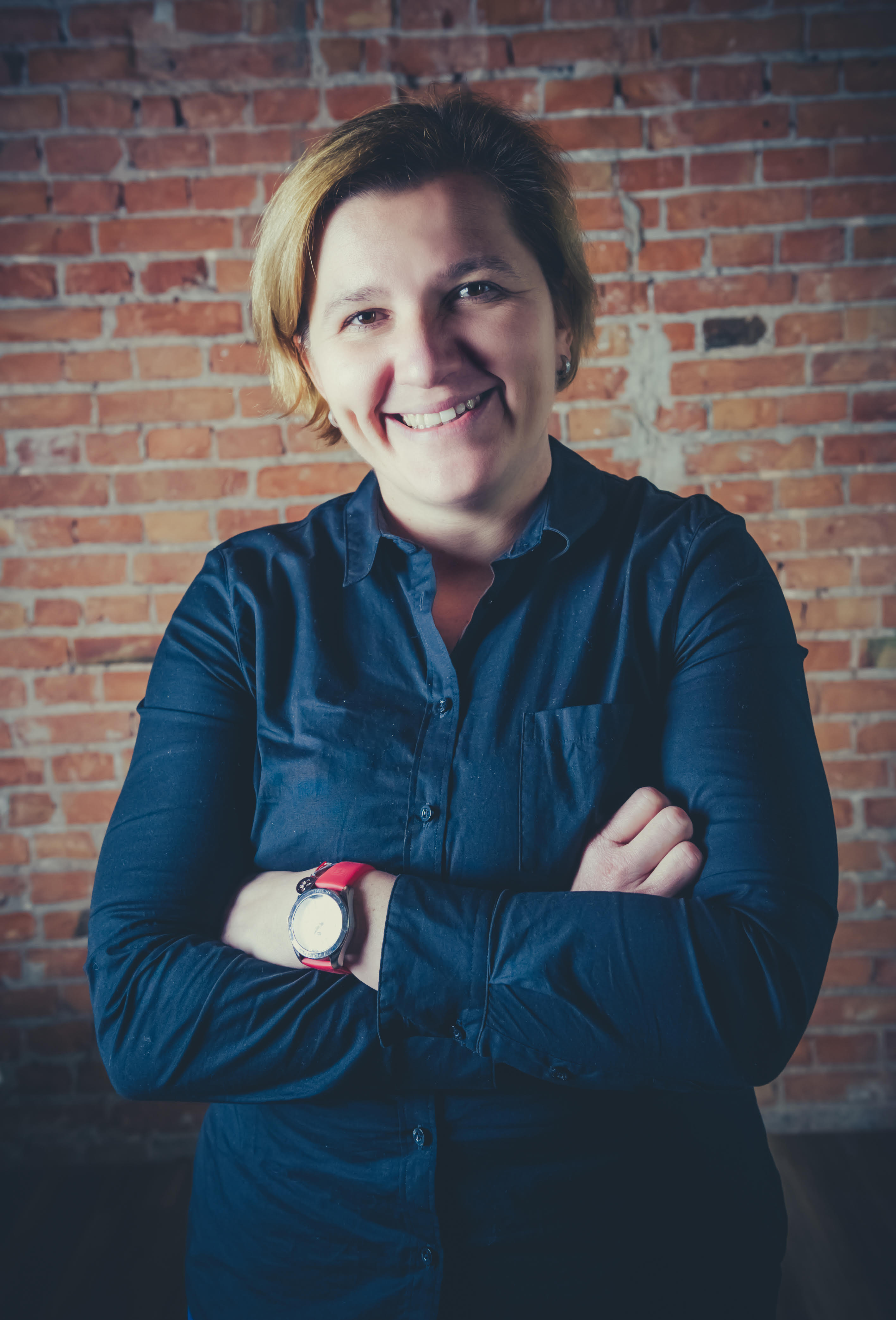 Uśmiechnięta kobieta kobieta o krótkich blond włosach stojąca z założonymi rękami. Jest ubrana w granatową koszulę z guzikami.