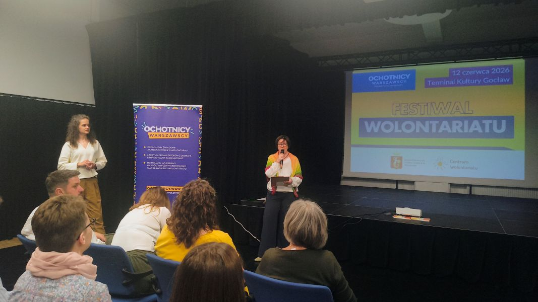 Kobieta przemawia do uczestników i uczestniczek konferencji. W tle ekran, na którym jest wyświetlony slajd z nazwą wydarzenia. 
