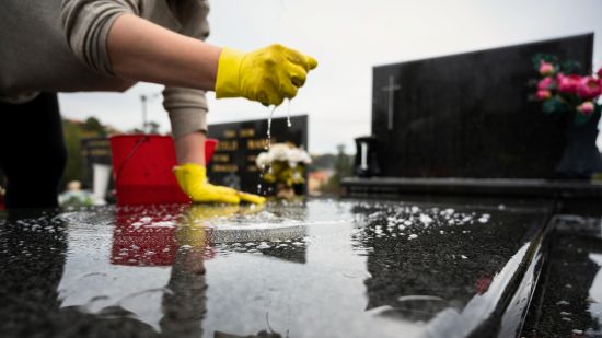 Ręce w żółtych rękawiczkach myją ciemną płytę nagrobną na cmentarzu, nagrobek polany wodą