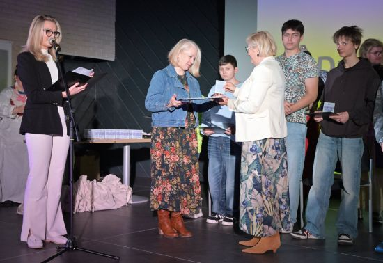 Elegancko ubrana kobieta przemawia przez mikrofon, obok inna kobieta składa drugiej gratulacje, w tle stojący młodzi ludzie