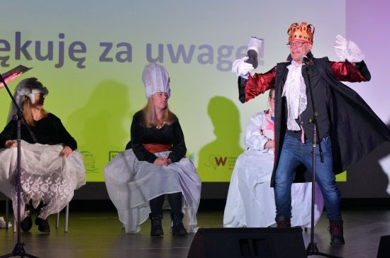 cztery osoby w strojach teatralnych na scenie, jedna z nich - mężczyzna z koroną na głowie szeroko gestykuluje i przemawia przez mikrofon, w tle napis z wyświetlonej prezentacji "Dziękuję za uwagę"