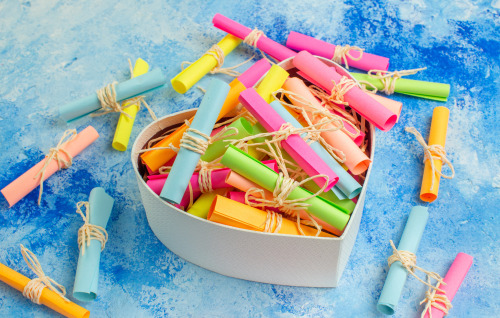 Zdjęcie pudełka w kształcie serca wypełnionego zwiniętymi w rulony kolorowymi karteczkami, przewiązanymi sznurkiem w kolorze naturalnym, tło w kolorze niebieskim