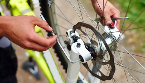 Zdjęcie przedstawiające proces przykręcania koła rowerowego, na zdjęciu fragment koła oraz dłonie trzymające klucze ampulowe