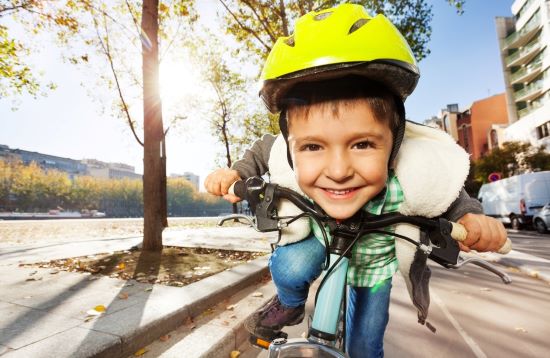 Zdjęcie uśmiechniętego kilkuletniego chłopca w cytrynowo żółtym kasku, jadącego pasem rowerowym wzdłuż ulicy, w tle słoneczny krajobraz miasta
