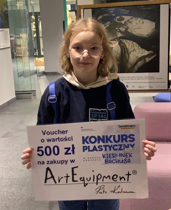 Uczennica w granatowej bluzie z logo SP 140 trzyma Voucher o wartości 500 zł jako nagrodę w konkursie plastycznym kierunek na Bachusa