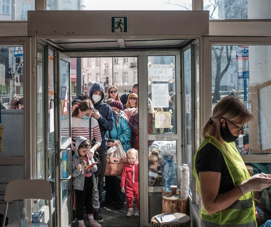 Grupa uchodźców z Ukrainy stojąca w drzwiach Centrum Pomocy Ukrainie. Wewnątrz wolontariuszka w żółtej kamizelce. 
