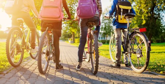 Zdjęcie grupy czterech młodych ludzi na rowerach, wykonane od tyłu, na zdjęciu widać tylne koła roweru, nogi oraz plecaki, góra zdjęcia jest przycięta, w tle chodnik, zieleń miejska i promienie słoneczne
