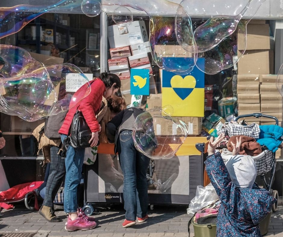 Trzy kobiety i dziecko przeglądają rzeczy przed budynkiem Centrum Pomocy Ukrainie. Nad dzieckiem duże bańki mydlane, obok wózek.
