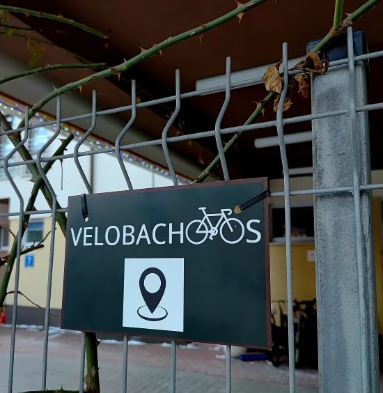 Tabliczka z napisem Velobachoos, symbolem roweru oraz pinezki zaznaczonej na mapie, przyczepiona do metalowej bramy wejściowej, bramę oplatają krzewy róż, w tle żółty budynek i krajobraz jesienno-zimowy