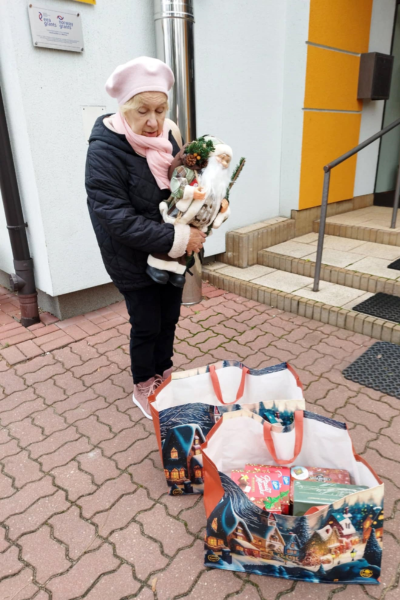 Kobieta stojąca przed budynkiem. W rękach trzyma figurkę Mikołaja. Obok stoją torby ze świątecznymi ozdobami i słodyczami.