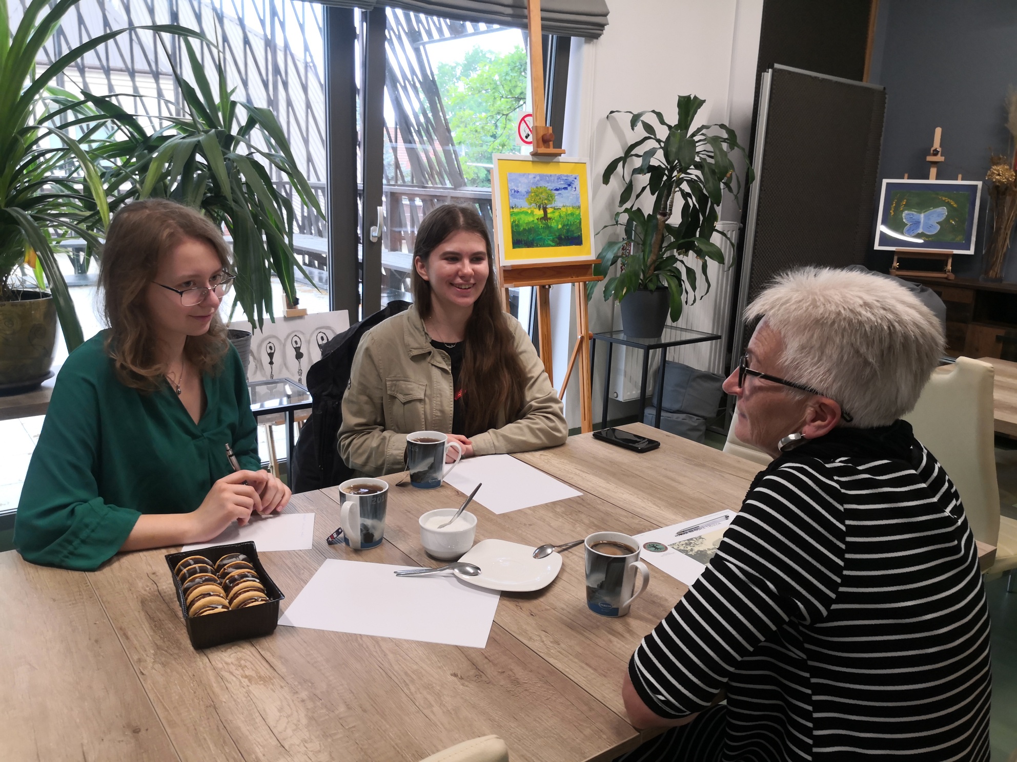 Dwie uśmiechnięte nastolatki siedzące naprzeciwko seniorki w krótkich włosach i bluzce w paski. Na stole między nimi znajdują się kubki z herbatą, paczka ciastek oraz kartki i długopisy.