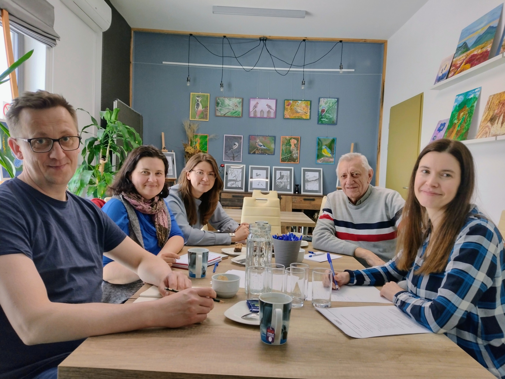 Trzy kobiety i dwóch mężczyzn siedzących przy stole i patrzących w na wprost w kamerę. Na ścianach w pomieszczeniu widać kolorowe obrazy przedstawiające florę i faunę.