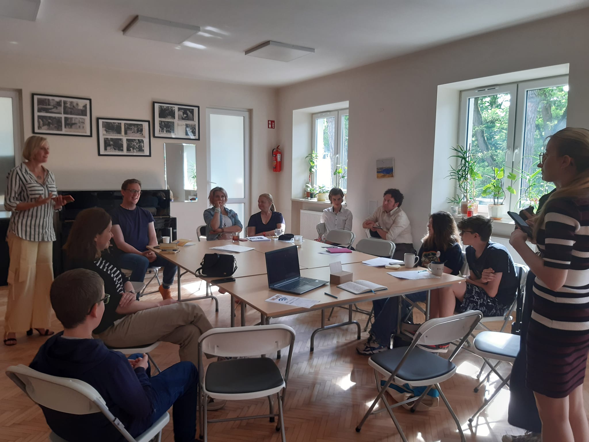 Grupa ludzi siedząca dookoła prostokątnego, brązowego stołu. U szczytu stołu stoją dwie kobiety. 