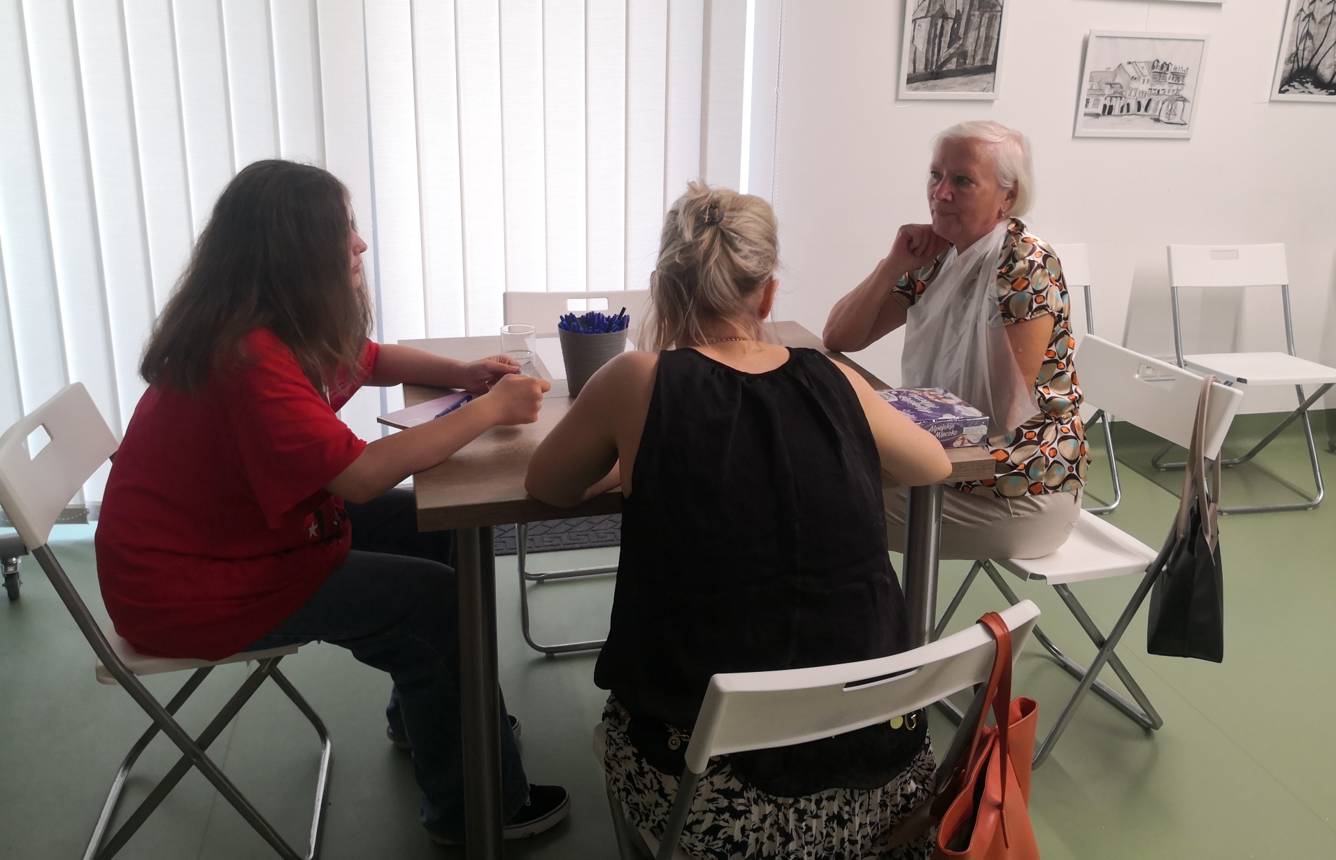 Trzy kobiety siedzące przy kwadratowym stole, na którym znajdują się kartki, długopisy oraz paczka ciastek. 