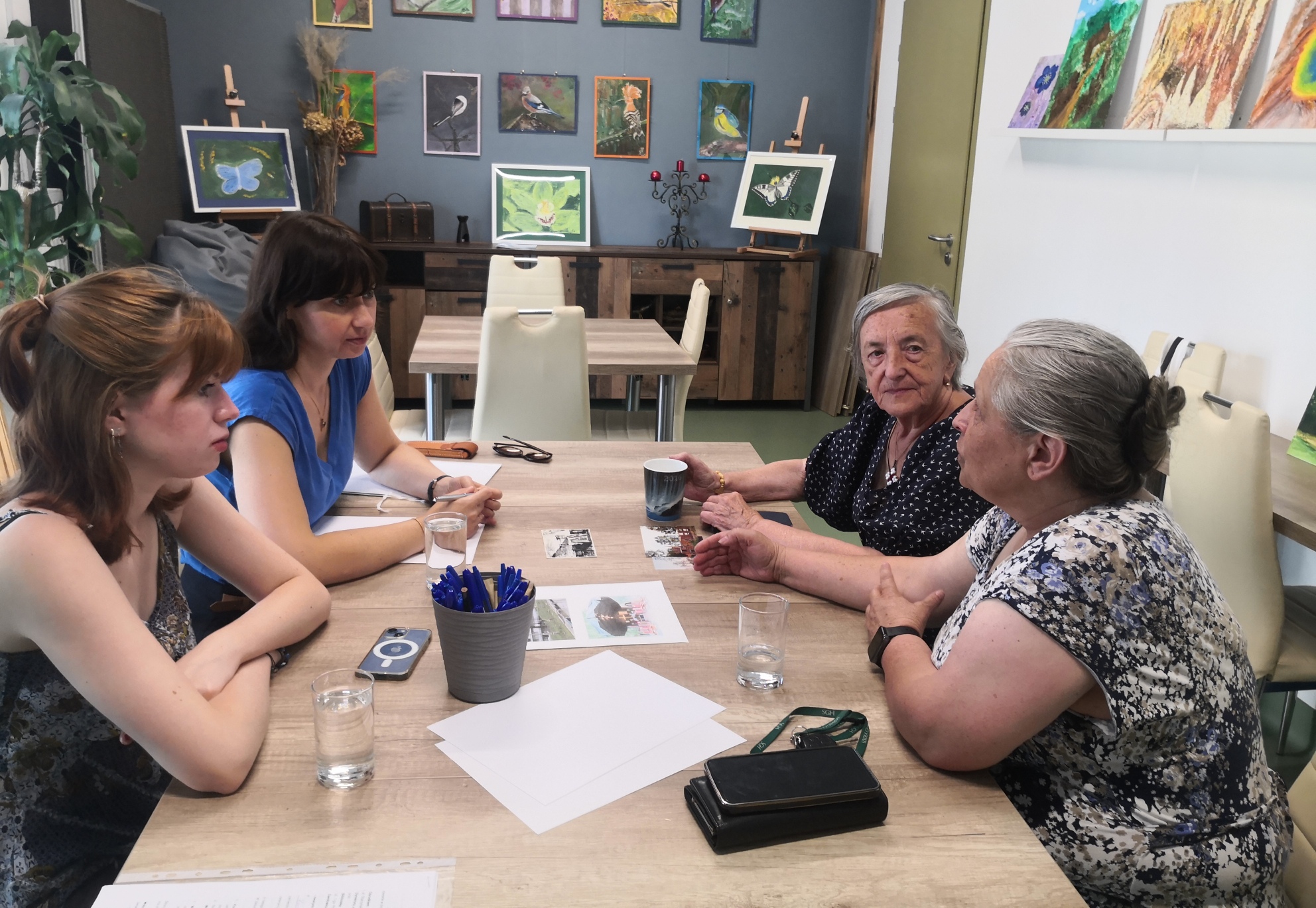 Cztery kobiety siedzące przy stole, na którym rozłożone są kartki, długopisy oraz szklanki z wodą. Trzy kobiety wpatrują się w czwartą - seniorkę wskazującą na zdjęcie leżące na stole.