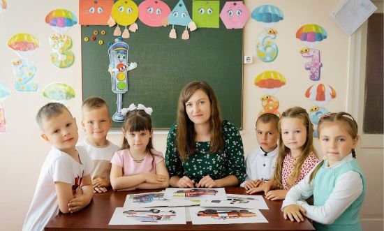 sześcioro dzieci w wieku wczesnoszkolnym stoi koło nauczycielki, uśmiechają się, w tle zielona tablica szkolna, nad tablicą kolorowe kartki z różnymi minami oraz kolorowe cyfry z parasolkami z papieru