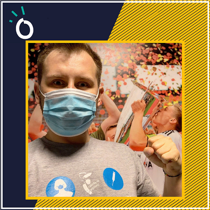 Łukasz Kachnowicz w maseczce i koszulce wolontariusza na tle tapety z piłkarzem Legii unoszącym puchar