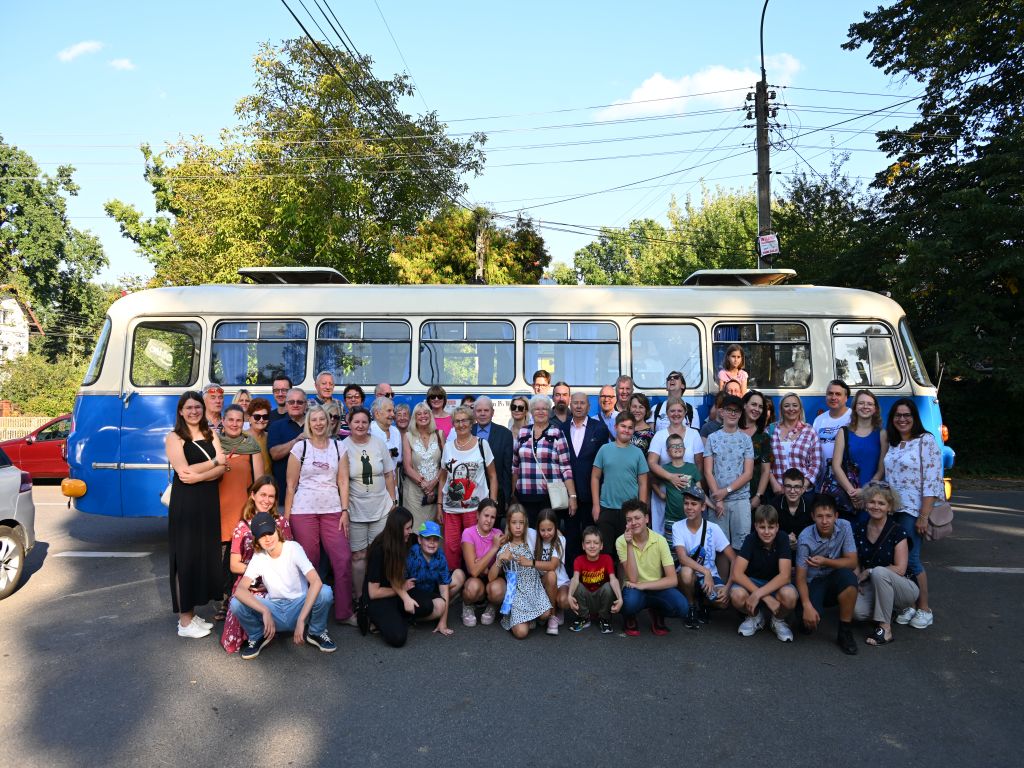 Grupa ludzi stojąca przed niebieskim samochodem typu "ogórek" i uśmiechająca się do kamery.
