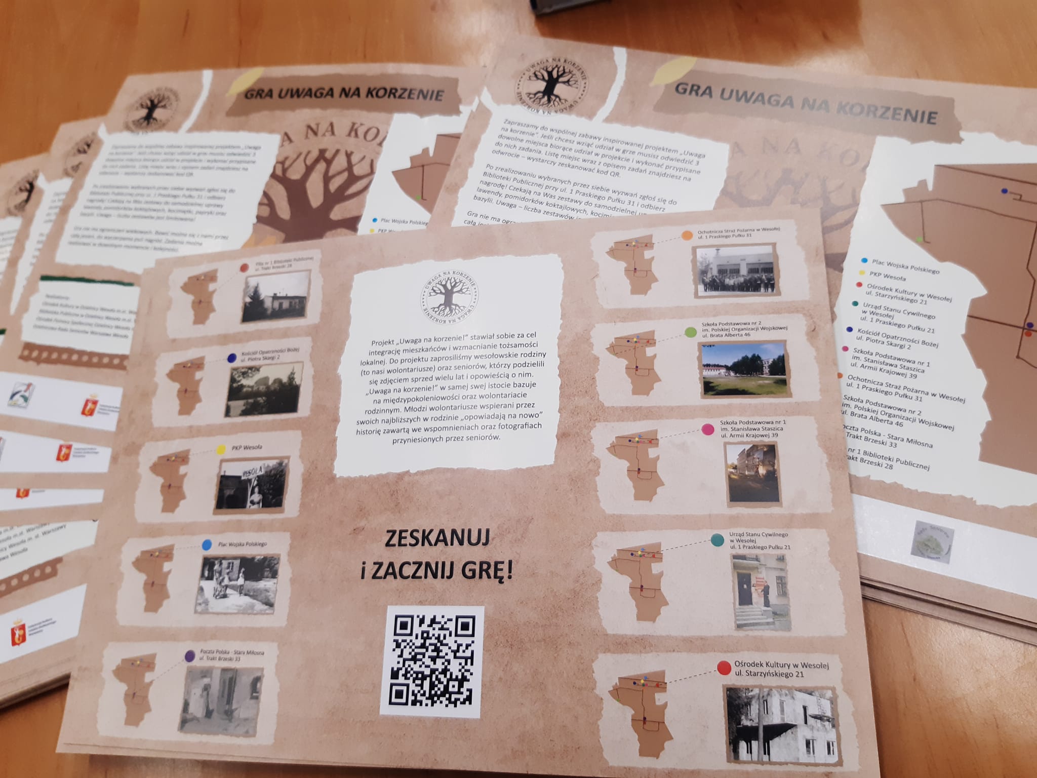 Stos plansz "uwaga na korzenie" z fragmentami map, zdjęciami oraz kodem QR i napisem "zeskanuj i zacznij grę" 