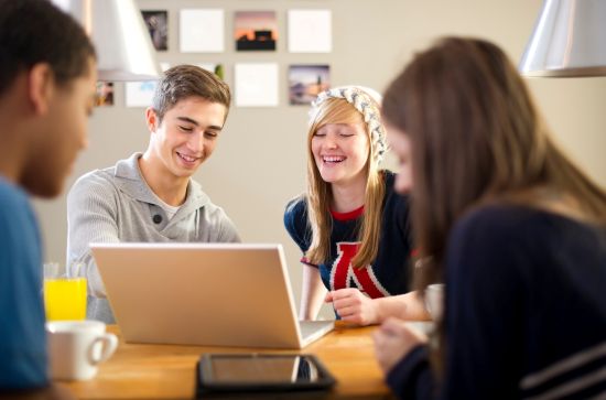 Czwórka młodych ludzi siedzących przy jednym laptopie, rozmawiających i śmiejących się