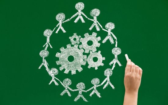 Zielona tablica i narysowane kredą symkboliczne postacie stojące w kręgu, w środku kręgu narysowane również białą kredą koła zębate