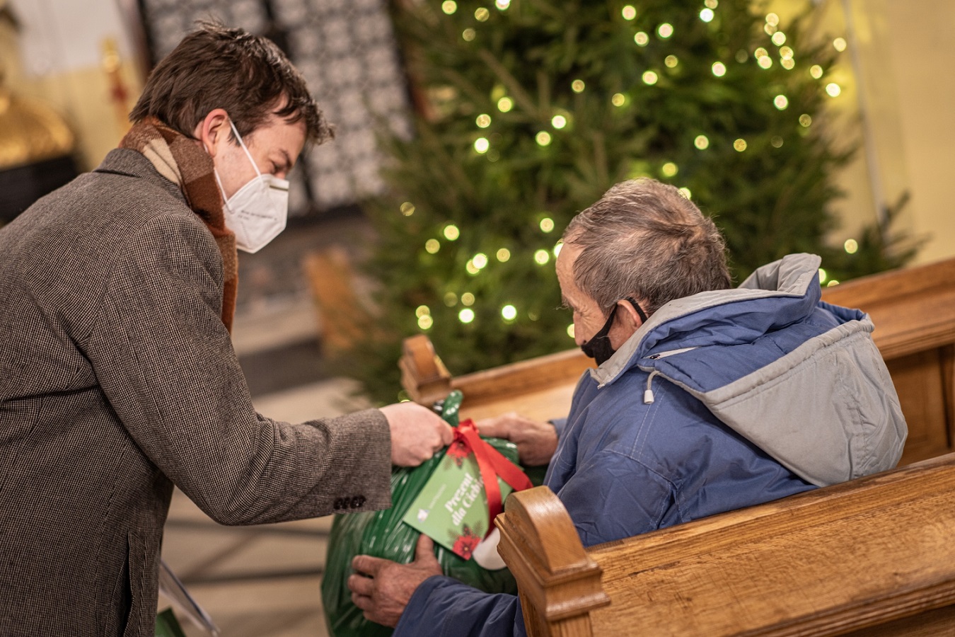 Wolontariusz przekazuje paczkę osobie bezdomnej siedzącej w ławie kościelnej. W tle choinka.