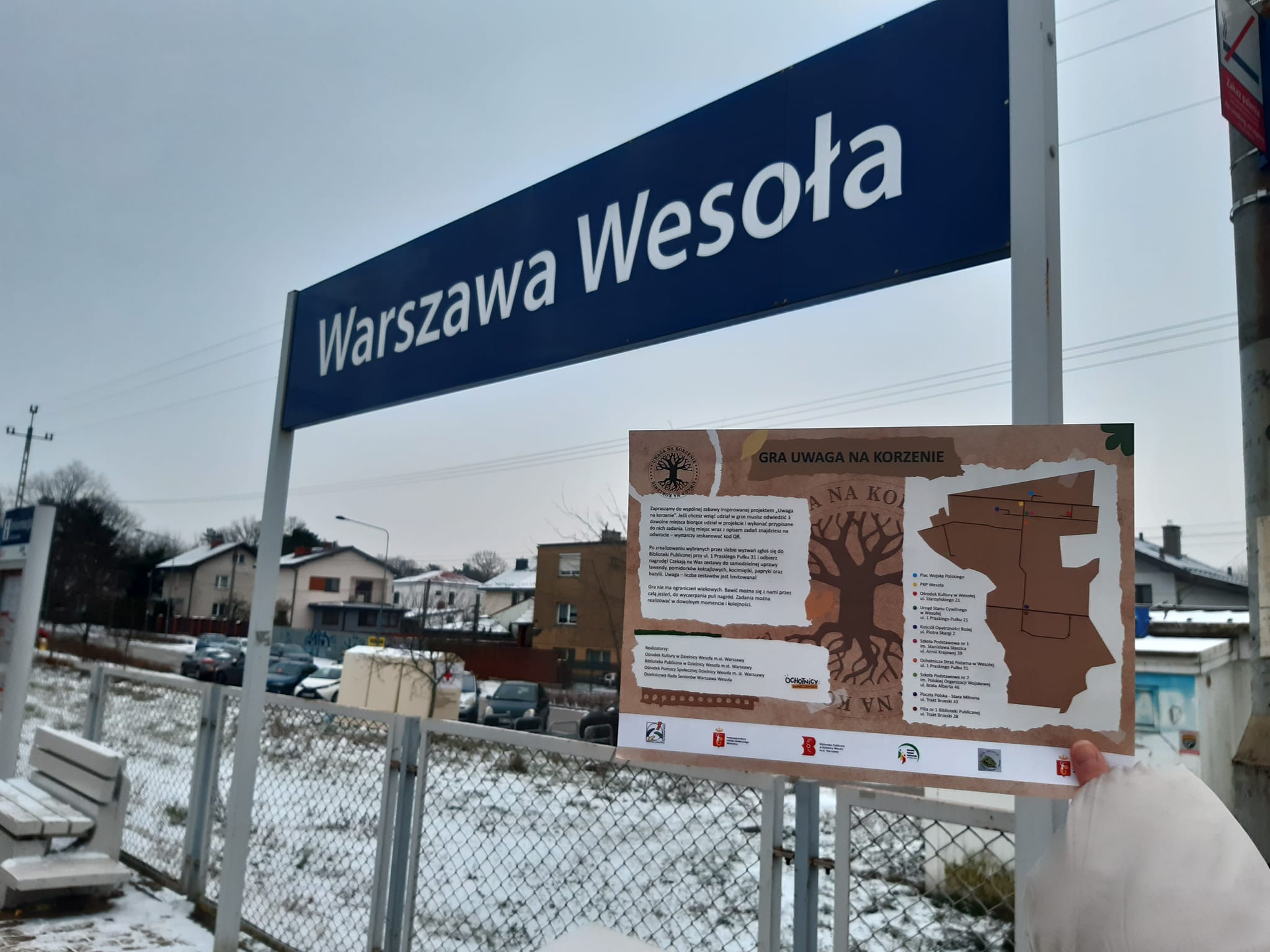 Plakat "uwaga na korzenie" na tle tablicy na stacji kolejowej "Warszawa Wesoła"