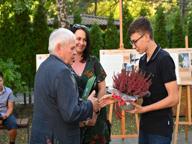 Senior, chłopiec i kobieta stojący na placu i trzymający zieloną teczkę i doniczkę z kwiatem. Chłopiec i senior ściskają swoje dłonie. W tle widać ustawione plansze z projektu "Uwaga na korzenie" 