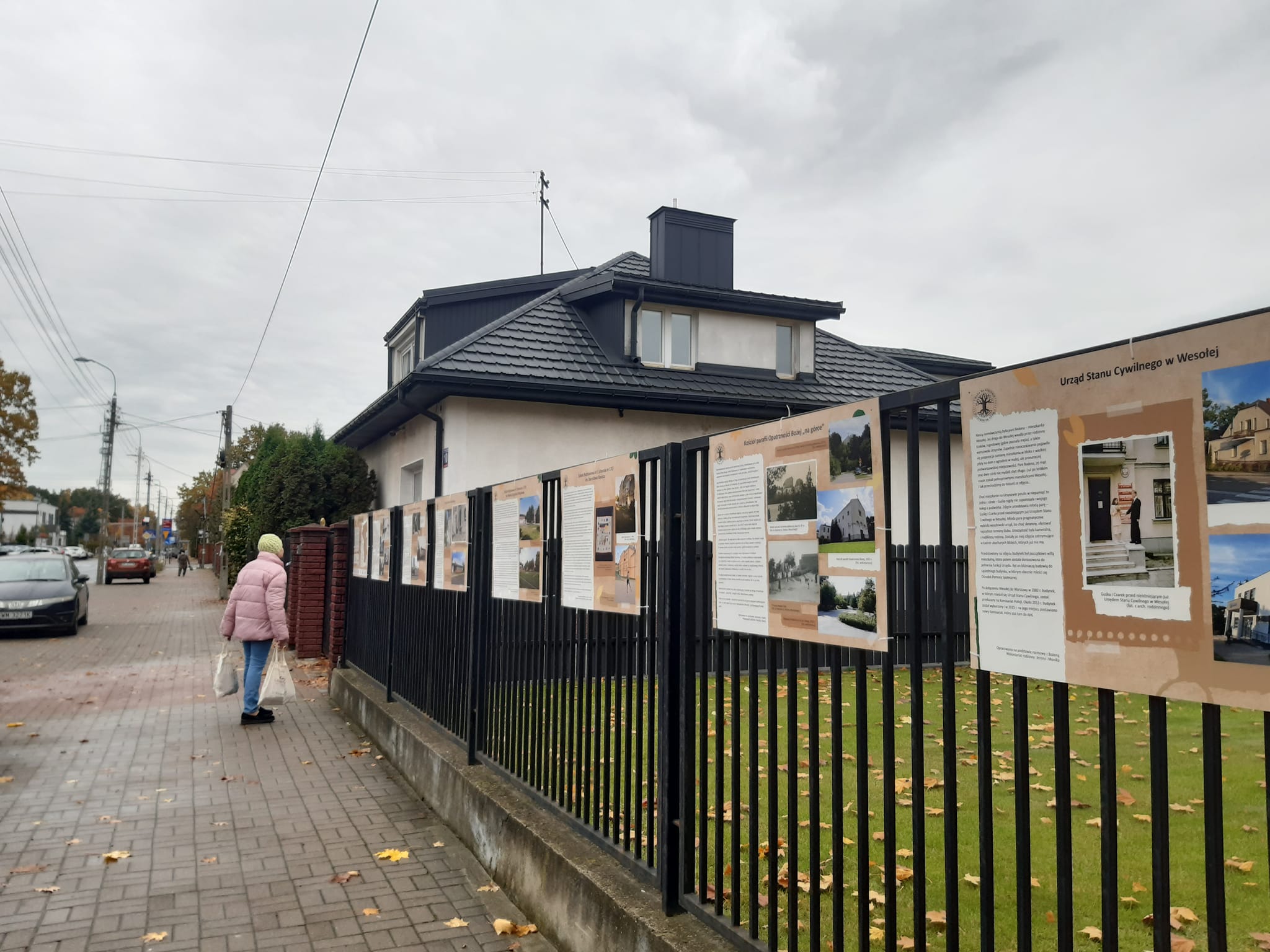 Plansze "Uwaga na korzenie" zawieszone na ogrodzeniu. W tle widać sylwetkę osoby w różowej kurtce oglądającej plansze. 