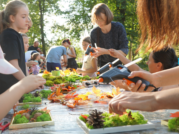 Grupa dorosłych i dzieci przy stole wykonuje prace artystyczne z użyciem szyszek, mchu, kamieni, liści. W tle widać drzewa.