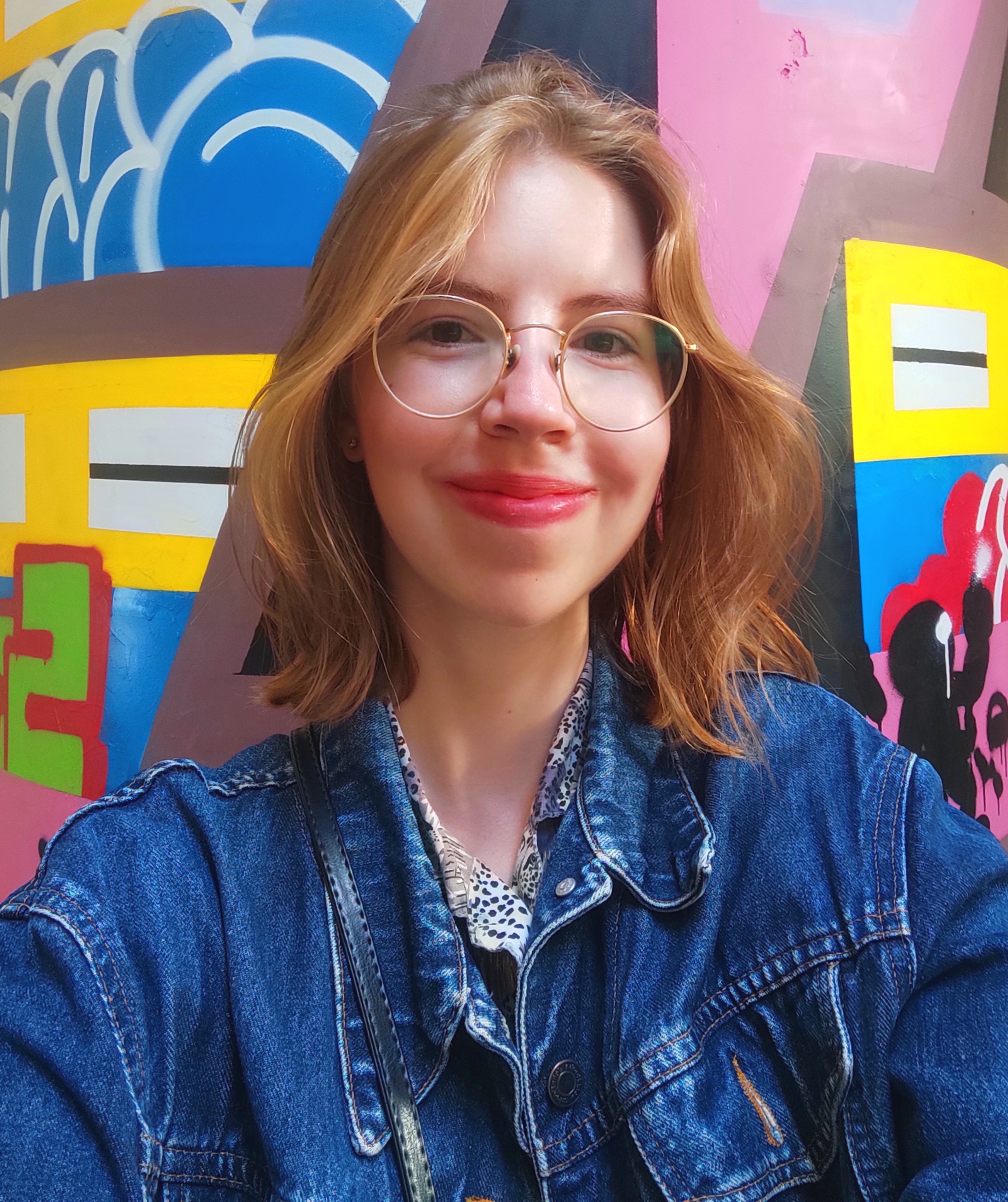 Zdjęcie portretowe wolontariuszki Mariki. Ma rude falowane włosy do ramion i okrągłe okulary w złotych oprawkach. Jest ubrana w dżinsową kurtkę i stoi na tle kolorowego muralu.