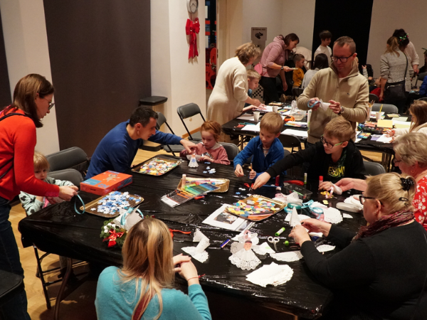 Pomieszczenie, w którym ustawione są dwa stoły. Osoby w różnym wieku stoją bądź siedzą przy stołach, wykonując prace plastyczne. 