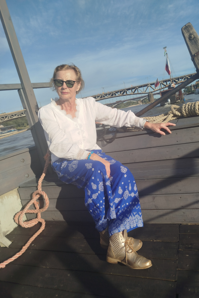 Pani Hanna siedząca na barce. W tle Wisła. Na twarzy ma okulary przeciwsłoneczne. Nosi białą koszulę, długie, niebieskie i zwiewne spodnie, złote buty za kostkę.