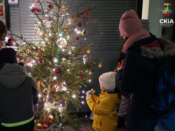Choinka z ozdobami świątecznymi, obok niej stoją osoby ubrane w kurtki, czapki i szaliki