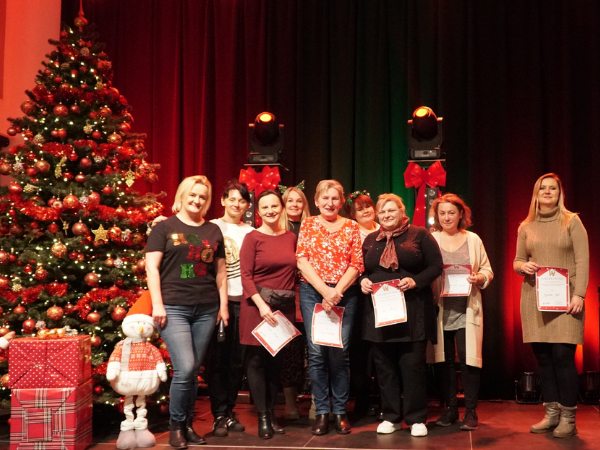 Scena, na której znajduje się choinka, prezenty i świąteczne ozdoby. Na środku niej stoi grupa osób, które trzymają w dłoniach kartki