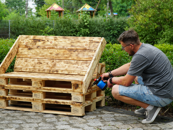 Mężczyzna kucający przy drewnianej ławce i trzymający dozownik z brązową farbą do malowania.