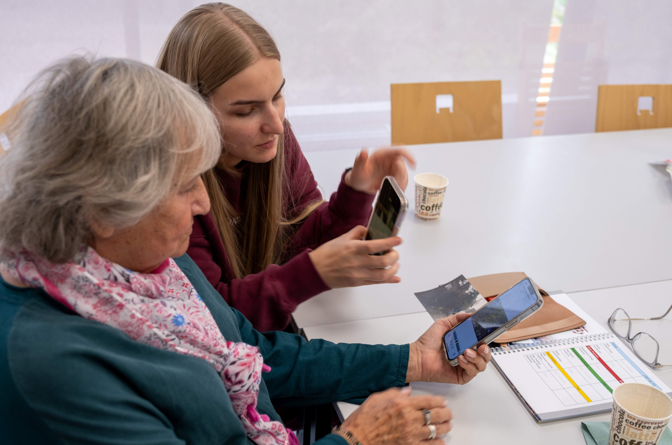Seniorka i młodsza uczestniczka siedzą przy białym stole, wspólnie przeglądając zdjęcia na smartfonach.