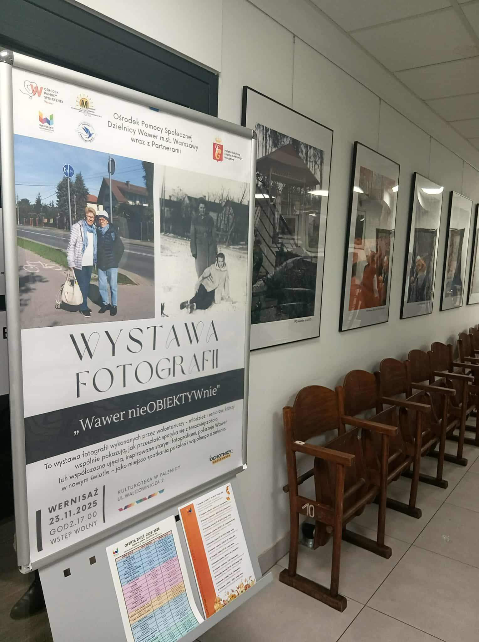 Plakat wystawy fotograficznej „Wawer nieOBIEKTYwnie” stojący w korytarzu przed rzędem drewnianych krzeseł i zawieszonymi na ścianie zdjęciami.