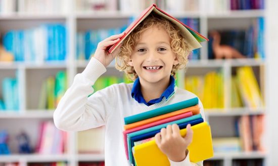 Chłopiec z rozłożoną książką na głowie i stosem książek w ręce