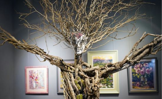 Rzeźba człowieka z rozłożonymi rękami, wykonana z gałęzi drzewa, w tle wystawa obrazów