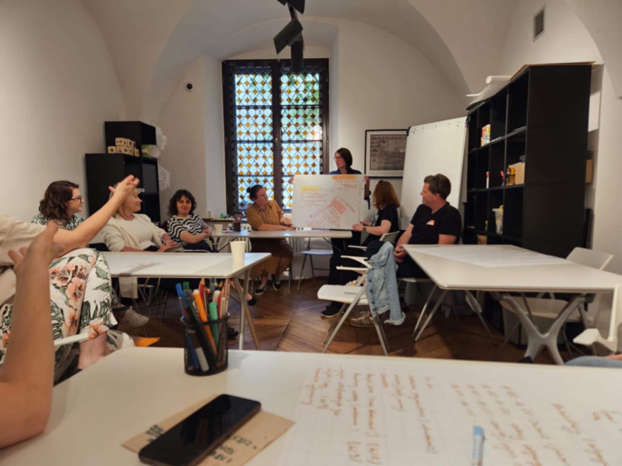 Koordynatorki i koordynatorzy wolontariatu z warszawskich muzeów w sali warsztatowej Muzeum Warszawy. Jedna osoba omawia wypracowane pomysły przy flipcharcie, pozostałe osoby siedzą i patrzą w jej stronę. 